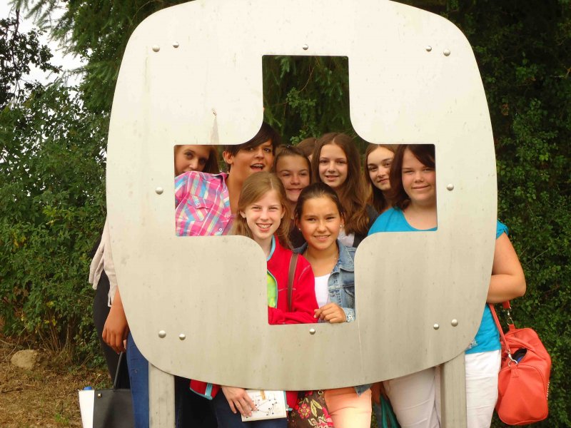 Eine Gruppe Schülerinnen steht im Freien hinter einer großen Metallskulptur und schaut lachend durch deren Ausschnitt.