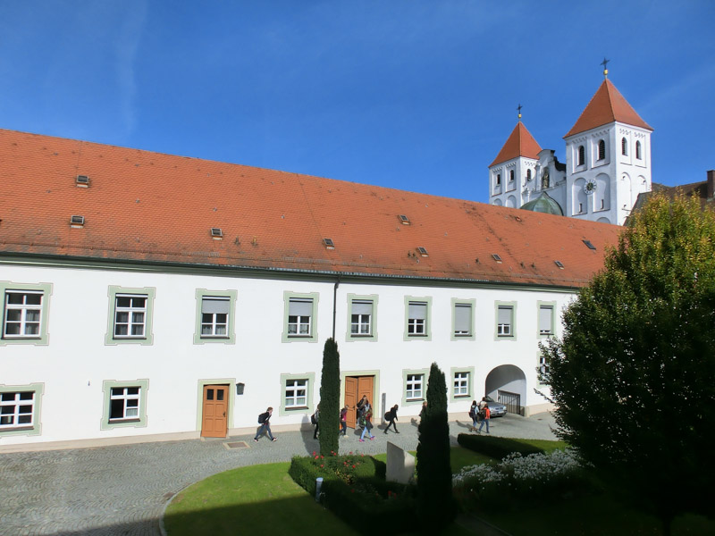 Der langgestreckte Schultrakt mit rotem Ziegeldach, dahinter die beiden Türme der Mutterhauskirche. Über den Innenhof gehen Schülerinnen.