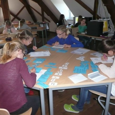 Schülerinnen sitzen um einen großen Tisch und ordnen beschriftete Karten. Im Hintergrund arbeiten weitere an Einzeltischen unter der Dachschräge.